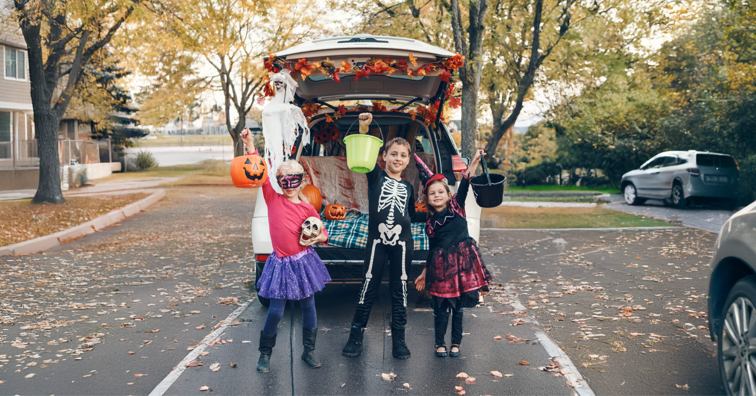 Three children in Halloween costumes stand in front of a car decorated for a trunk-or-treat event, holding candy buckets on a leaf-covered street.