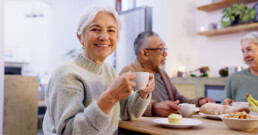 A smiling older woman holds a coffee mug at a table with two other seniors
