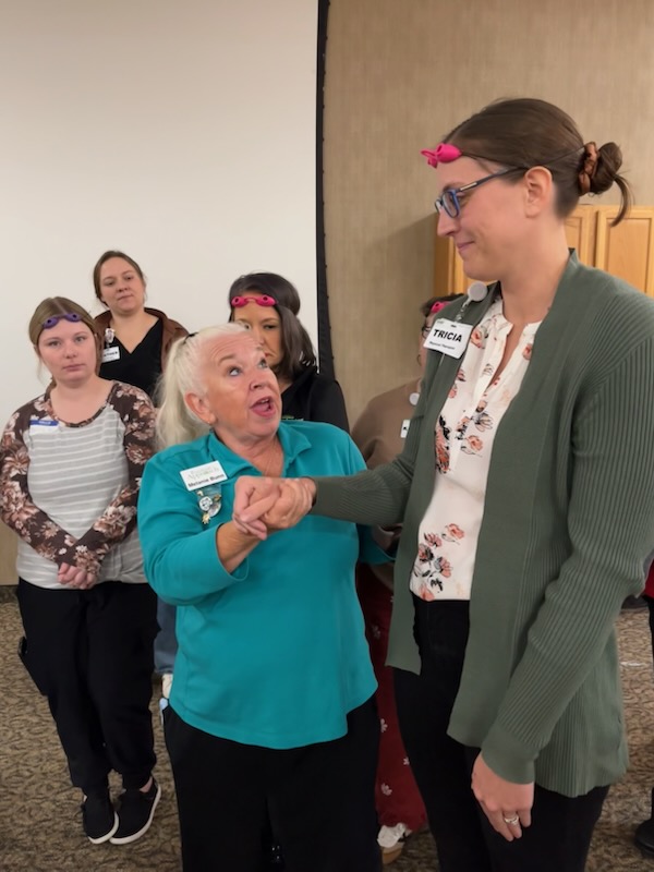 A woman in a teal shirt demonstrates a hand-holding technique to a taller woman named Tricia, while three other women observe in a conference room.
