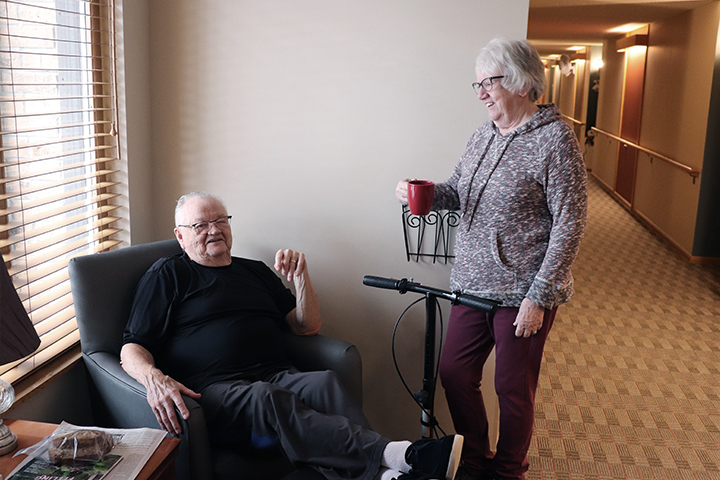 A woman with short-cropped hair and eye glasses brings a cup of coffee to her husband who's sitting in a lounge chair with his feet up on a transport scooter.
