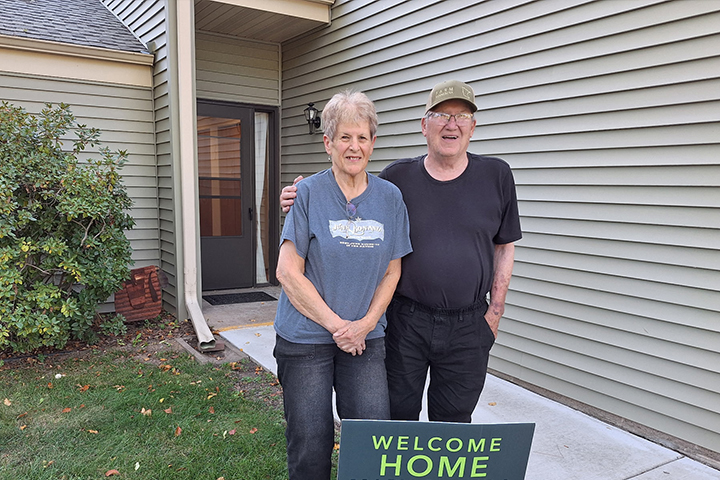 An smiling older couple stands in front of their townhome next to a sign that reads "Welcome Home". He has his arm around her shoulder.