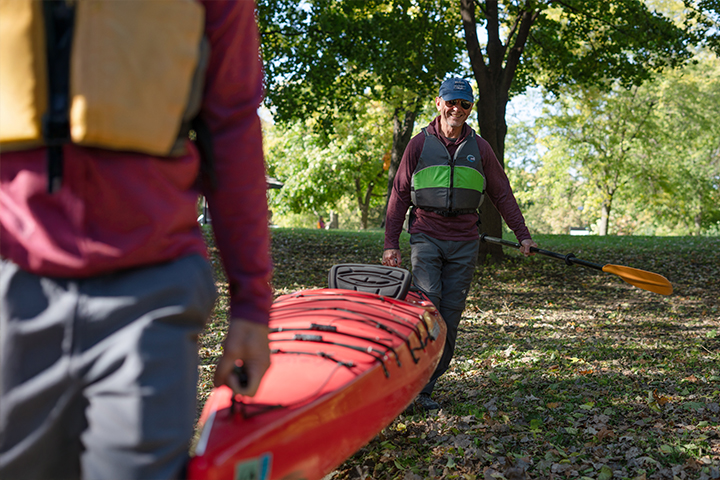 An older man wearing a cap and life jacket holds a paddle in one hand and carries the back end of a red kayak with the other. Another person in the foreground carries the front end of the kayak.