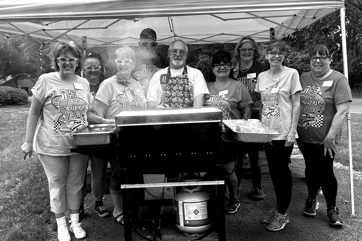 Black and white photo of female and male team members wearing themed t-shirts as they gather outside behind a gas grill during an event.