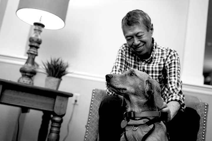 Black and white photo of a smiling older man sitting on a chair and petting a lab that sits at his feet.