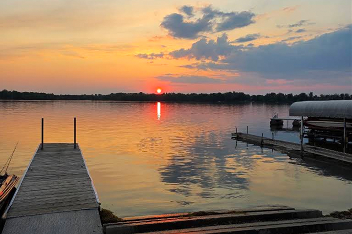 A beautiful sunset, multiple colors and a few clouds reflect off the lake with a dock and boat launch in the foreground.