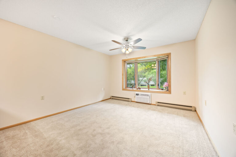 Interior apartment living room with tan carpeting, cream walls, ceiling fan, wall air conditioner unit, and a large window.