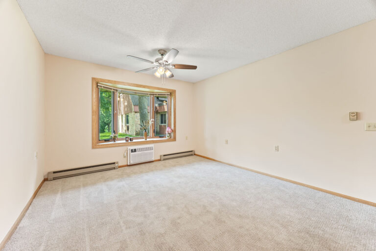 Interior apartment living room with tan carpeting, cream walls, ceiling fan, wall air conditioner unit, and a large window.