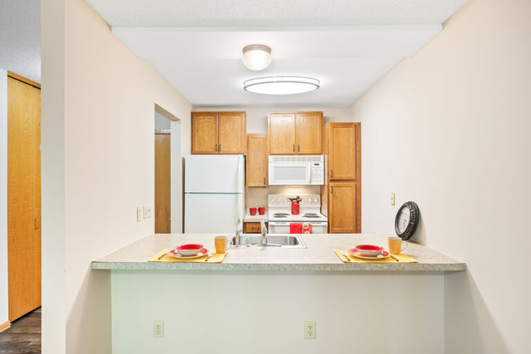 Interior apartment kitchen shown from dining area, featuring cream countertop bar with two place settings, stainless steel sink, and white appliances with wood cabinets.
