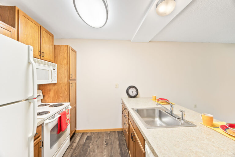 Interior apartment kitchen featuring white appliances, wood cabinets and cream countertop with stainless steel sink.