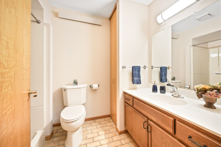Interior apartment bathroom featuring tile floor, white vanity, toilet and step-in shower, with wood cabinets.