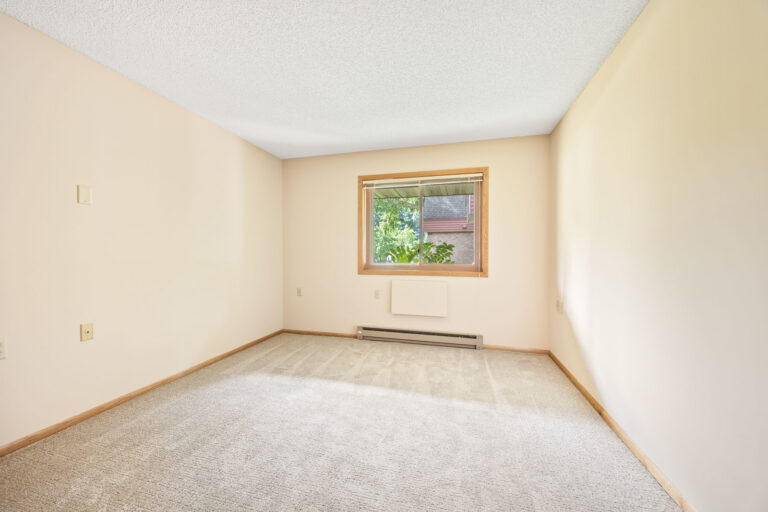 Interior apartment bedroom with tan carpeting, cream walls, and large window.