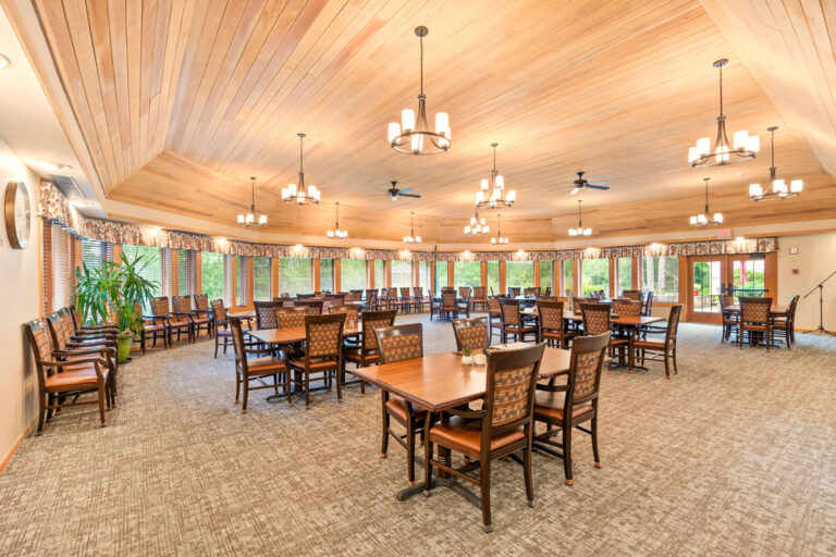 Interior large dining room, featuring carpeting, four-up tables with upholstered chairs, wood paneled ceiling, a wall of windows, and a double door connecting to an outdoor patio.