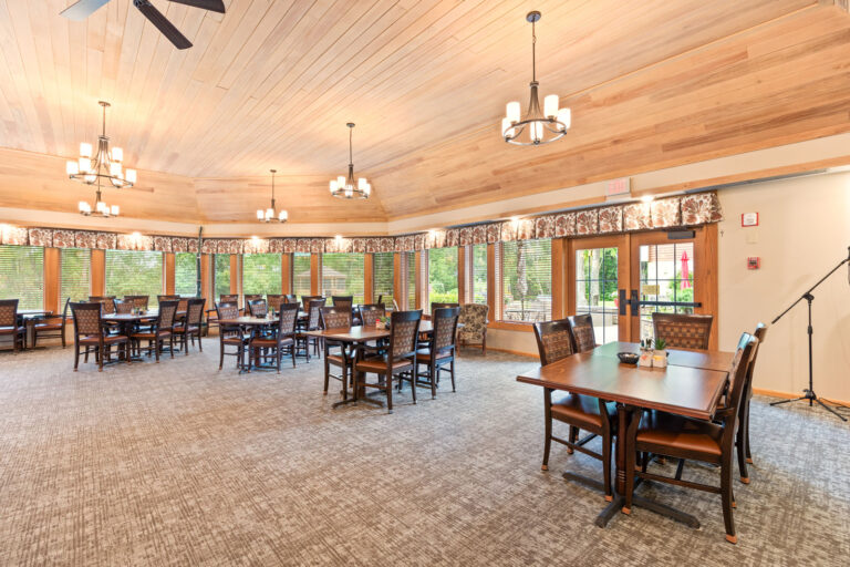 Interior large dining room, featuring carpeting, four-up tables with upholstered chairs, wood paneled ceiling, a wall of windows, and a double door connecting to an outdoor patio.