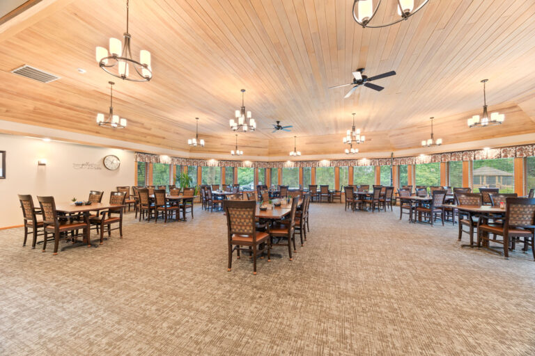 Interior large dining room, featuring carpeting, four-up tables with upholstered chairs, wood paneled ceiling, and a wall of windows letting in lots of natural light.