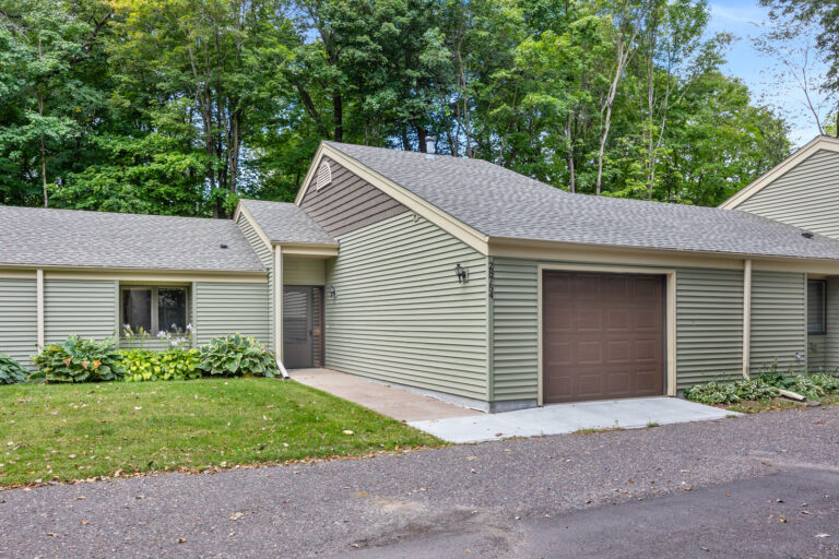 Exterior of a townhouse, featuring sage green siding, cream trim, and tan pitched roof. A sidewalk leads to the front door from the drive way and along the one-car garage with dark brown door.