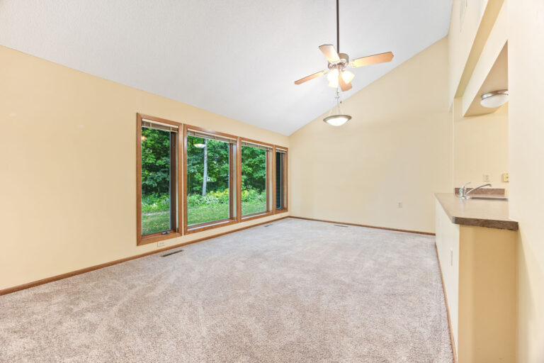 Interior living space with tan carpeting, cream walls, wood trim, vaulted ceiling with fan, and big windows with blinds.