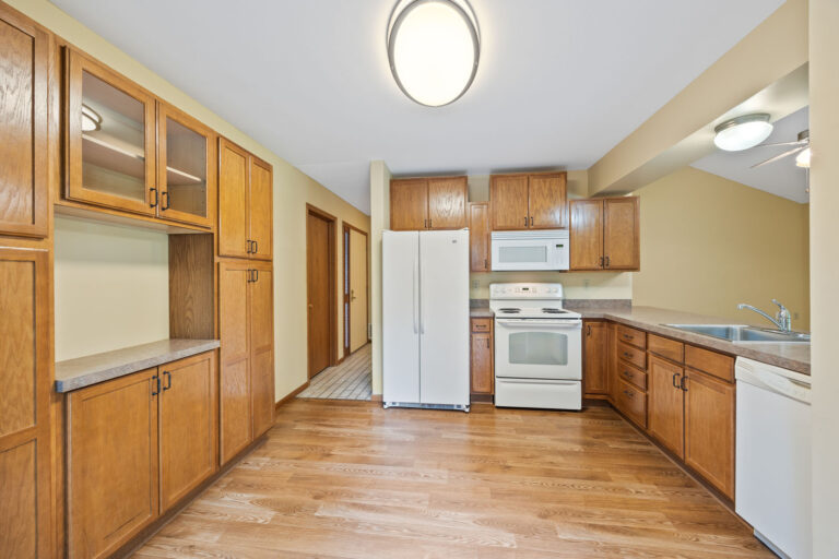 Interior kitchen with white appliances, wood cabinets, hardwood floor, and tiled hallway leading to entryway.