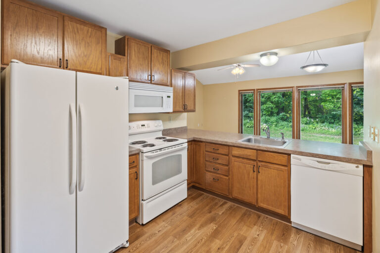 Interior kitchen with white appliances, wood cabinets, hardwood floor, and countertop open to living space.