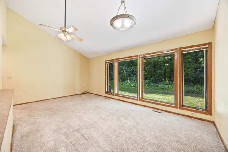 Interior living space with tan carpeting, cream walls, wood trim, vaulted ceiling with fan, and big windows with blinds.