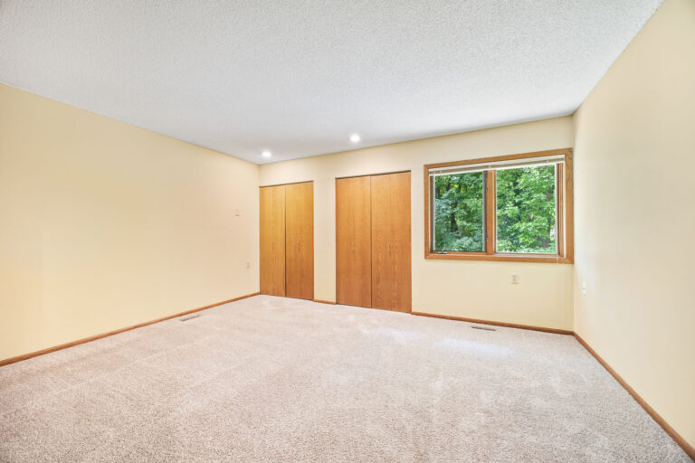 Interior bedroom with tan carpeting, cream walls, wood trim, two closets, and big windows with blinds.