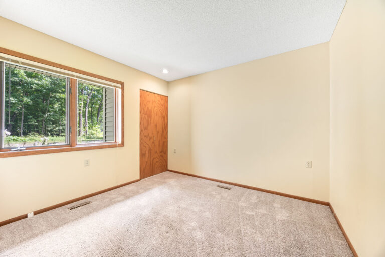 Interior bedroom with tan carpeting, cream walls, wood trim, closet, and big windows with blinds.