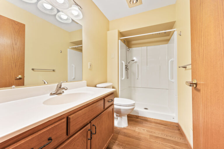 Interior bathroom with white vanity, toilet and step-in shower with wood cabinetry and cream walls.