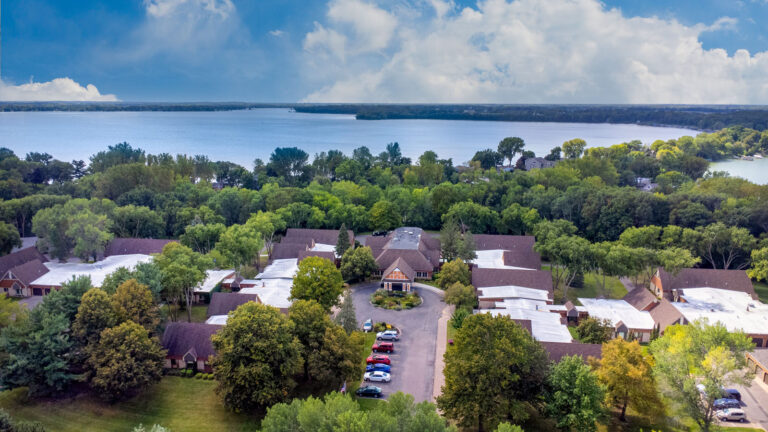 Southwest-facing aerial view of campus main entrance, featuring mature trees and proximity to two lakes separated by a peninsula. Blue sky and clouds meet the horizon.
