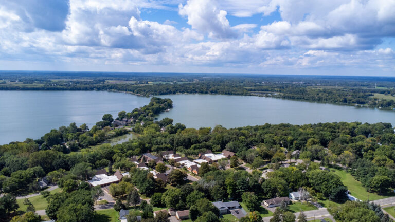 West-facing aerial view of campus, featuring mature trees and proximity to two lakes separated by a peninsula. Blue sky and clouds meet the horizon.