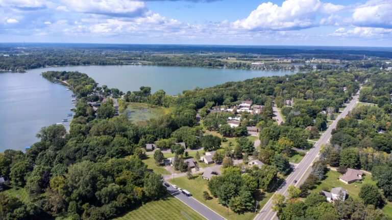 Northwest-facing aerial view of campus, featuring local road, mature trees and proximity to two lakes separated by a large peninsula. Blue sky and clouds stretch to the horizon.