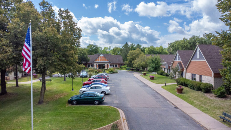 A driveway with cars parked perpendicular on one side leads passed pitched-roof apartment buildings to the main entrance and car port, featuring terracotta, beige and white Scandinavian design. An American flag flies on a flag pole in the foreground.