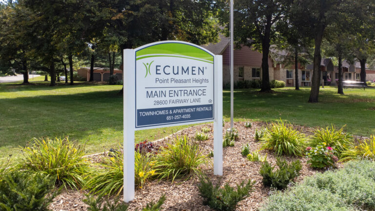 A sign surrounded by landscaping mulch and shrubs reads "Ecumen Point Pleasant Heights Main Entrance".