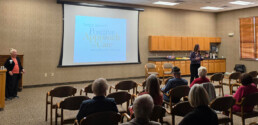 A woman presents to an audience in a conference room with a projection screen displaying the Teepa Snow Positive Approach to Care brand logo.
