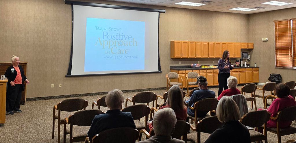 A woman presents to an audience in a conference room with a projection screen displaying the Teepa Snow Positive Approach to Care brand logo.