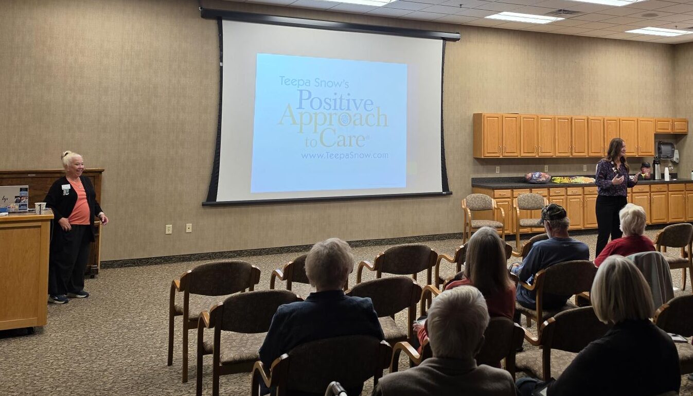 A woman presents to an audience in a conference room with a projection screen displaying the Teepa Snow Positive Approach to Care brand logo.